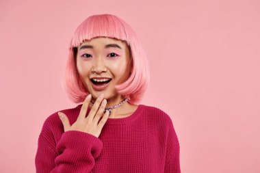 happy asian girl in vibrant sweater with pearl necklace expressing admiration on pink background clipart