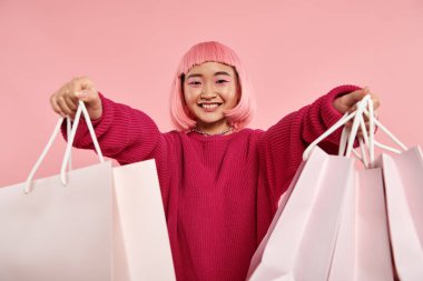 Güzel Asyalı genç kız geniş bir gülümsemeyle pembe arka planda alışveriş çantalarını tutuyor.