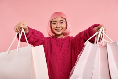 beautiful asian young girl smiling broadly and holding out to shopping bags on pink background clipart