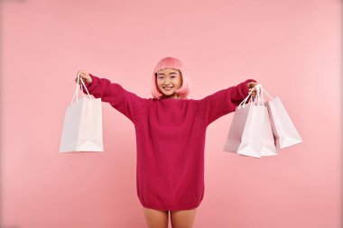 Gülümseyen ve pembe arka planda alışveriş torbaları tutan neşeli Asyalı genç kadın.