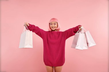 cheerful asian young woman smiling and holding shopping bags on pink background clipart