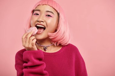 portrait of cheerful asian woman with pink hair eating mochi with admiration on vibrant background clipart