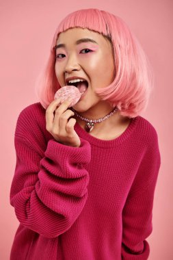 Pembe saçlı ve makyajlı sevimli Asyalı genç kız canlı arka planda mochi yiyor.