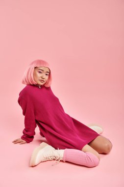 20 'li yaşlarda, pembe saçlı, büyük süveterli çekici bir kadın rahat bir şekilde arka planda oturuyor.