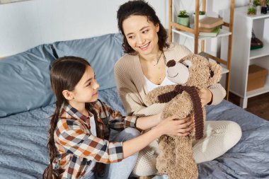Bir anne, sıcak bir yatakta kızının yanında otururken nazikçe oyuncak ayıyı tutar..