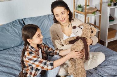 A mother tenderly holds a teddy bear while sitting next to her daughter on a cozy bed. clipart