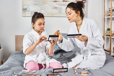 Esmer bir anne ve kızının beyaz banyo cübbesiyle bir yatakta oturduğu huzurlu bir sahne..