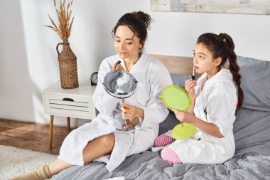 Esmer bir anne ve kızı, ikisi de beyaz banyo bornozu giymiş, dudak parlatıcısı sürüyor.
