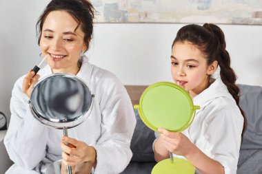 Beyaz banyo bornozu giymiş esmer bir anne ve kızı, dudak parlatıcısı sürüyorlar, rahat bir yatakta birlikte oturuyorlar..