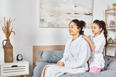 İki kadın, bir anne ve kız, ikisi de kumral saçlı, beyaz cüppeli bir yatakta, birlikte huzurlu bir anı paylaşıyorlar..