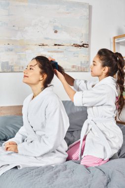 Beyaz banyo cübbesi giymiş esmer bir anne ve kız, rahat bir yatakta huzur içinde oturuyorlar..