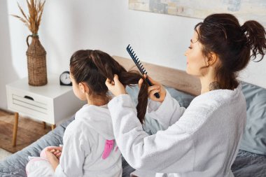 A brunette mother in a white bathrobe sits on a bed combing the hair of her young daughter, also in a white bathrobe. clipart