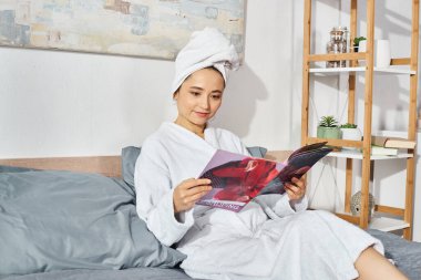 Beyaz banyo bornozu giymiş esmer bir kadın yatakta oturmuş, dergi okumaya dalmış.