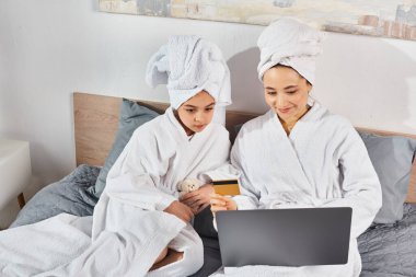 İki kadın, esmer bir anne ve kız, beyaz banyo bornozları içinde bir yatakta oturuyor, dizüstü bilgisayarla meşgul..