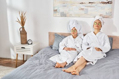 Beyaz banyo bornozu giymiş esmer bir anne ve kız kafalarına havlu sarılmış bir şekilde bir yatakta oturuyorlar..