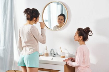 A brunette woman brushes her teeth in front of a mirror in a modern bathroom while her preteen daughter looks on. clipart