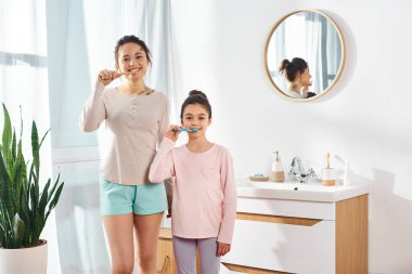 Esmer bir kadın ve küçük kızı, güzellik ve hijyen rutinlerinin bir parçası olarak modern bir banyoda dişlerini fırçalıyorlar..