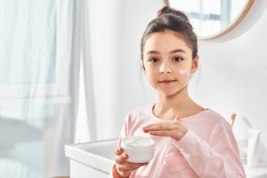 Siyah saçlı genç bir kız elinde bir kavanoz krem tutuyor, modern bir banyoda cilt bakımı ya da güzellik gösterisi yapıyor..
