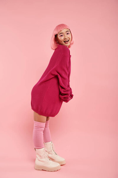 cheerful asian woman in big sweater outfit standing in sideways pose against pink background