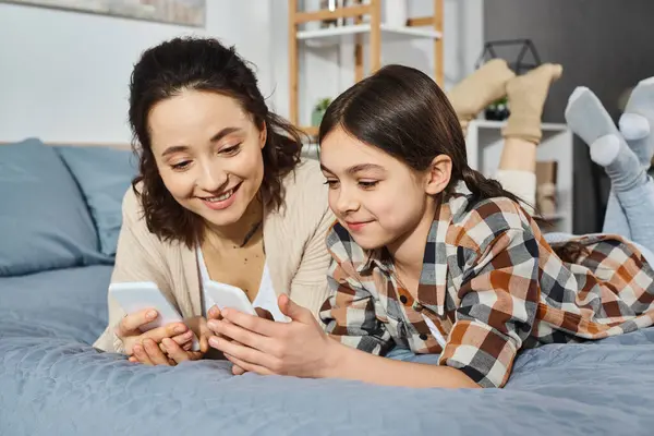 mother and daughter, lying on bed, focused on shared cell phone screen, enjoying quality time together.