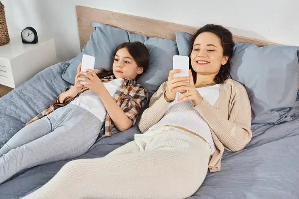 a mother and daughter, lying on a bed, holding up cell phones and bonding over their technology.