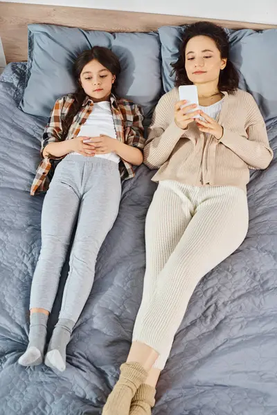 A mother and daughter are laying on a bed, absorbed in their smartphones, enjoying quality time together at home.