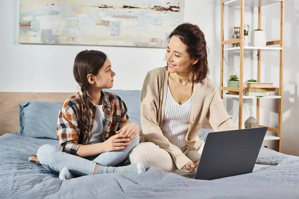 A mother and daughter sitting on a bed, engrossed in a laptop together.