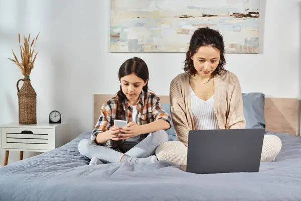 mother and daughter, sitting on a bed, focused on a laptop screen, sharing a special moment together at home.