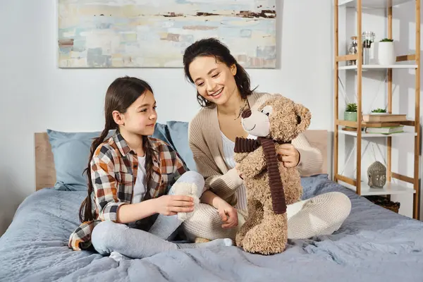 A mother and daughter sit on a bed, enjoying quality time with a teddy bear between them.