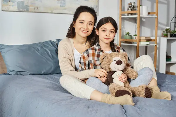 Two women, a mother and her daughter, sit on a bed with a teddy bear, sharing a moment of closeness and connection.