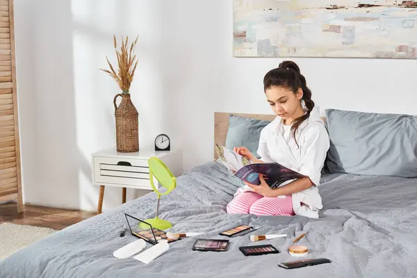 A serene scene of a brunette preteen girl in a white bath robe sitting on a bed, engrossed in a magazine with cosmetics nearby.