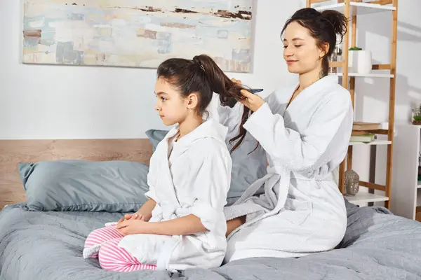 A brunette mother and daughter sit on a bed, wearing white bath robes, sharing a tender moment together.