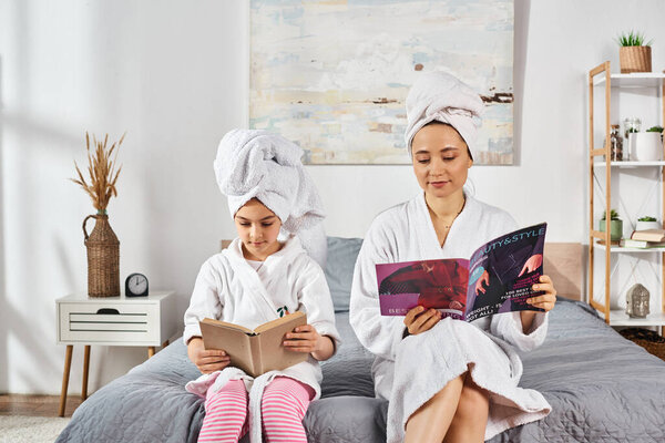 A brunette mother and daughter in white bath robes sit on a bed, immersed in a book and magazine