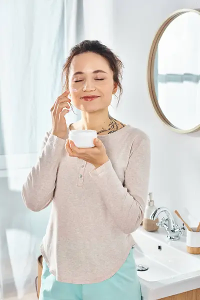 ブルネット女性は 美しさと衛生的な活動の中で エレガントなバスルームの設定でクリームと美容瓶を保持し 鏡で立っています — ストック写真
