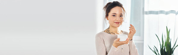 A brunette woman holds a jar of cream, gazing directly at the camera with a serene expression on her face.