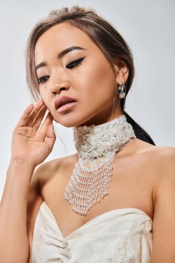 Zarif makyajlı güzel Asyalı kadın aşağı bakıyor ve açık arkaplanda yüzüne dokunuyor.