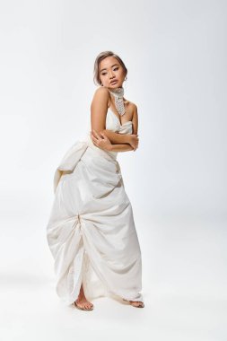 graceful asian young woman in white elegant dress hug herself and look to down on light background clipart