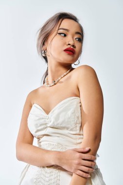 alluring asian woman in her 20s with red lips and pearl necklace look to side on white background clipart