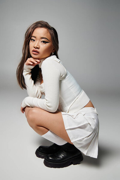 asian woman in white stylish outfit crouching down sideways against grey background