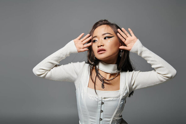 alluring asian woman in her 20s looking to side and posing with hands against grey background