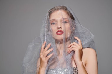 Genç bir kadın klasik bir güzellik sergiliyor, kafası narin bir peçeyle süslenmiş, gri bir arka planda sakin bir stüdyoda..