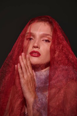 Kızıl saçlı bir kadın klasik güzellik yayar, gizemli bir cazibeyle yüzünü bir stüdyo ortamında örterek poz verir..