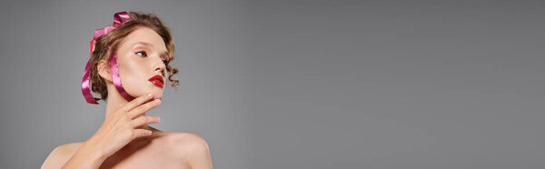 A young woman exudes classic beauty, posing in a studio with a pink bow on her head against a grey backdrop.