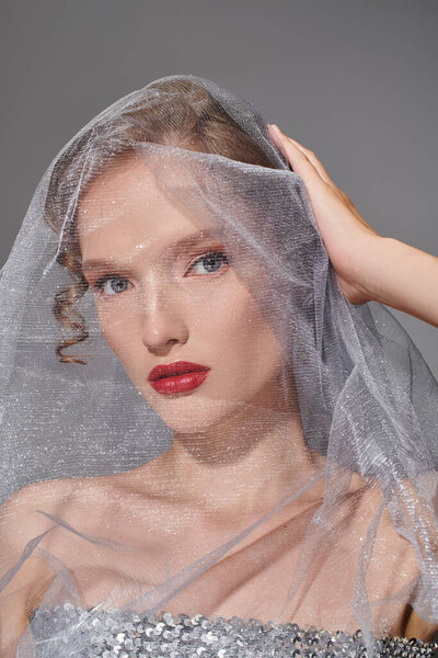 A young woman exudes classic beauty as she poses with a veil draped delicately over her head in a studio setting.