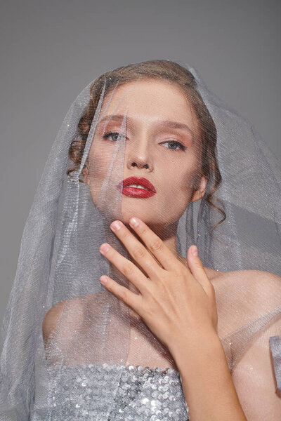 A young woman exuding classic beauty, wearing a veil and vibrant red lipstick in a studio setting on a grey background.