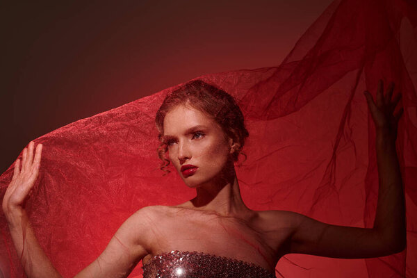 A young woman exudes classic beauty in a strapless dress, holding a vibrant red cloth while striking a pose in a studio setting on a black background.