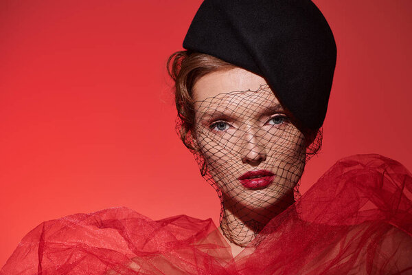 A young woman exudes classic beauty in a striking red dress and black hat as she poses confidently in a studio setting.