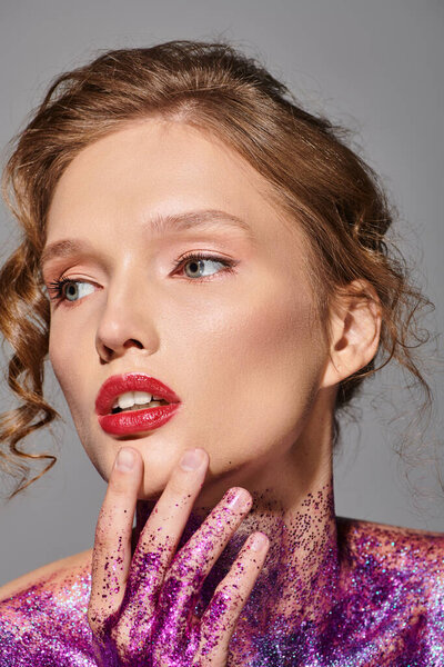 A young woman in a purple dress stands with her hands on her face, exuding classic beauty in a studio setting with a grey backdrop.