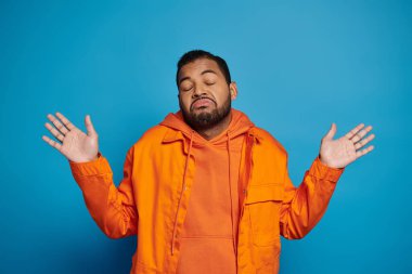 charismatic african american man in orange outfit showing that knows nothing on blue background clipart