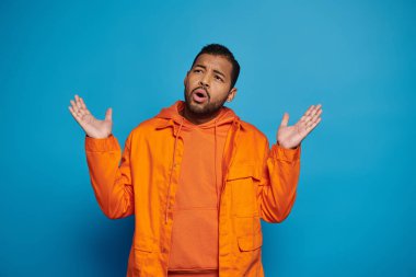 Kafası karışmış turuncu elbiseli Afrikalı Amerikalı adam mavi arka planda ellerini yana koyuyor.